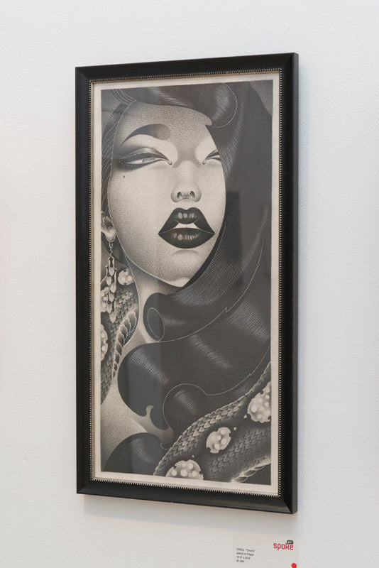 A stylized black-and-white framed portrait by ONEQ, titled Orochi, depicts a woman with bold makeup, large earrings, and flowing hair featuring intricate designs. Created in pencil on paper, it is displayed on a white wall.