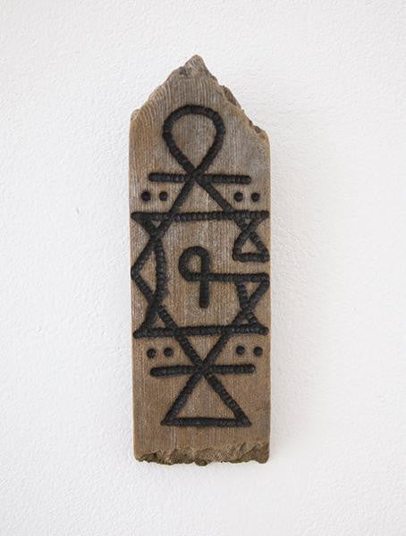 This GATS wooden plaque features carved geometric patterns—triangles, circles, and a top curved loop. Its central area showcases a stylized P. Crafted from burnt driftwood for a rustic look, its mounted on a white wall.
