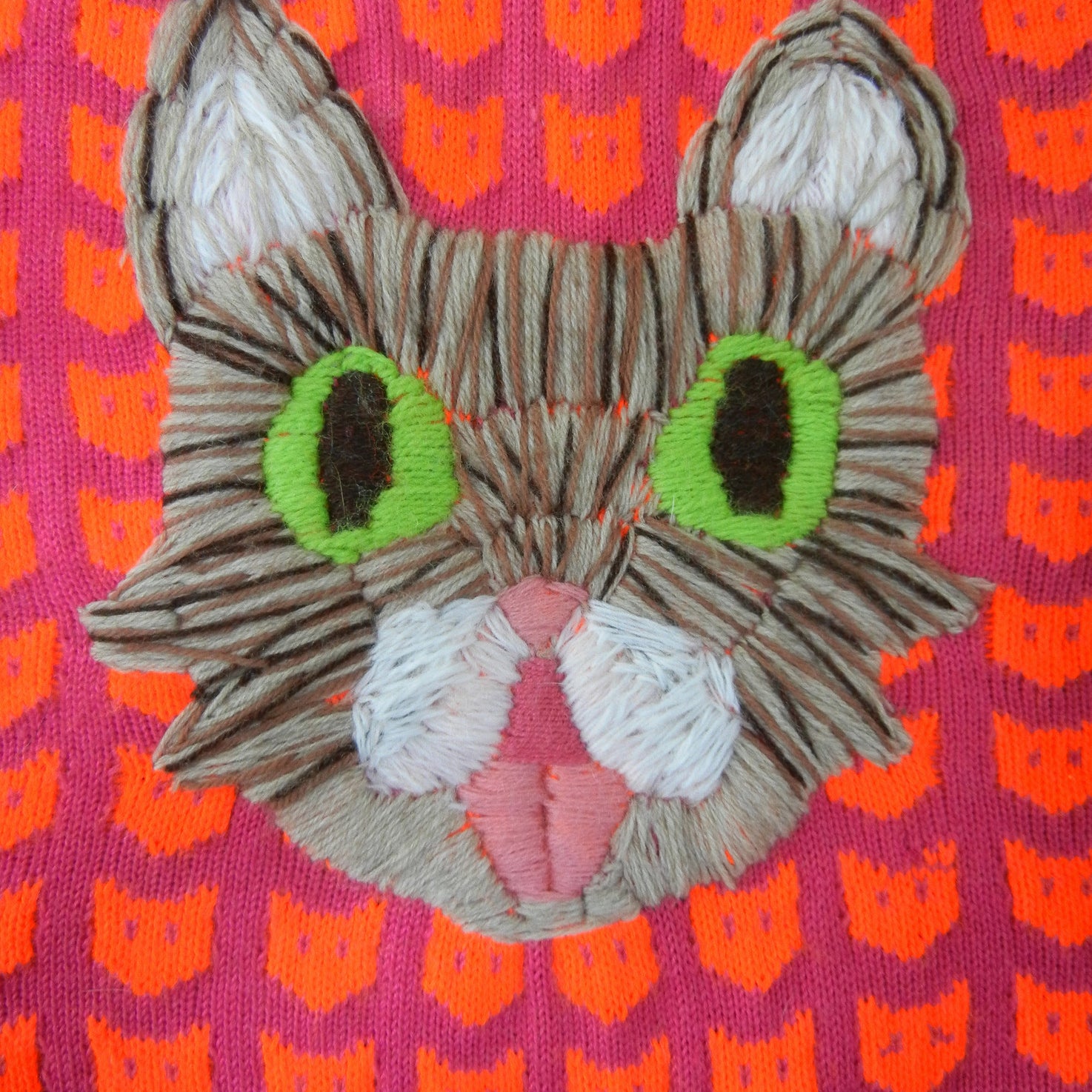 A vibrant Ishknits embroidery of a cats face, akin to Lil Bub, with green eyes, a pink nose, and tongue over a pink background with orange cats is perfect for display at San Franciscos Spoke Art gallery.
