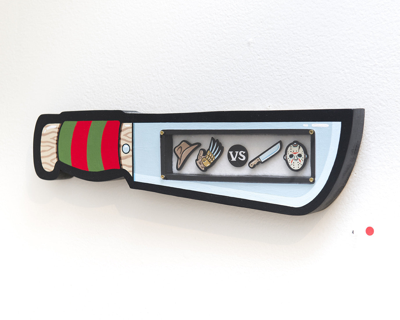 Crafted by Matt Ritchie, this wood-cut decorative knife features a striped handle and intricate blade designs, including a fedora, clawed glove, VS, machete, and hockey mask. It references Freddy vs. Jason and is accented with a small red dot on the right side against a white background.