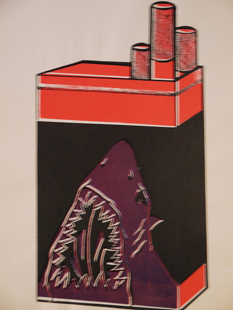 A Shark Toof cigarette pack illustration shows three cigarettes protruding, highlighted by a shark graphic with an open mouth for a warning. The hand-printed design in red, black, and white adds an edgy flair.