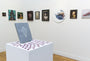 The gallerys interior features diverse framed paintings, including an Alex Louisa oil piece. In the foreground, a gray book with a geometric design rests on a white pedestal, surrounded by overlapping cards.