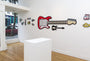 The Matt Ritchie art gallery features vibrant cartoon-style pieces on white walls, including a large red electric guitar, hand-cut wood items, and miniatures like a camera and car. A central pedestal complements the eclectic scene, with a window offering a street view.