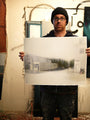 In a studio filled with paint splatters and supplies, a person in a beanie and glasses holds Brett Amorys limited edition painting depicting a blurred urban scene.