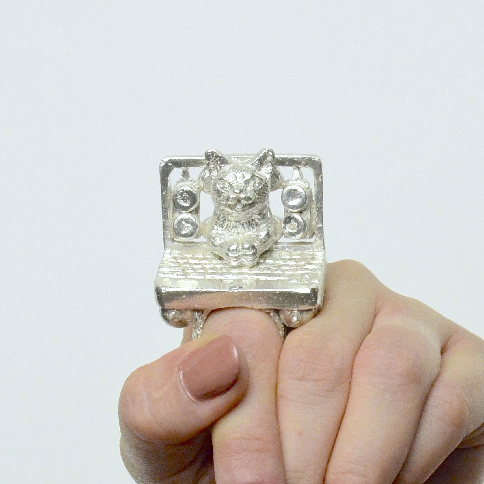 With painted nails, a person wears a Rebecca Rose Sterling Silver Ring featuring a detailed cat figurine sitting on a computer with speakers, echoing the whimsical charm of a San Francisco gallerys Lil Bub art exhibit.