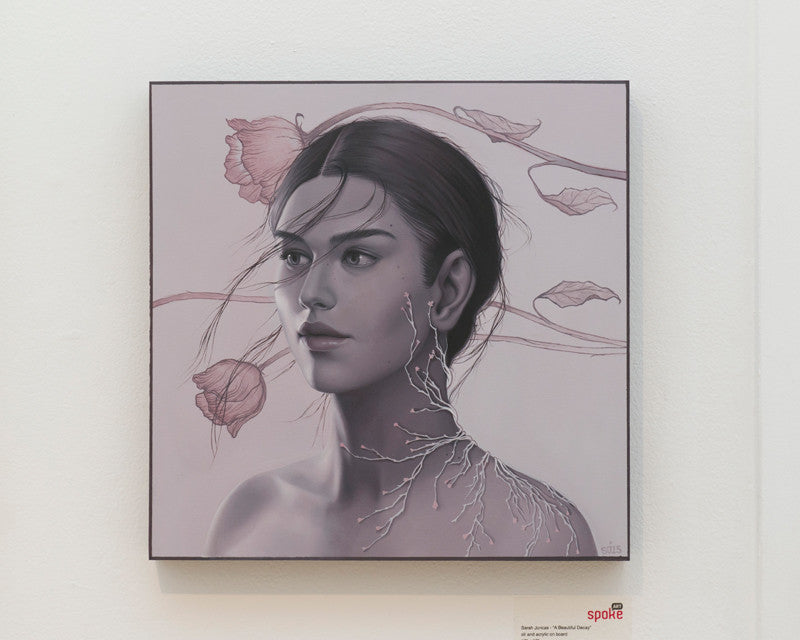 A Beautiful Decay by Sarah Joncas is a square painting with a woman of dark hair encircled by delicate flowers and vines. The piece uses monochromatic tones with hints of pink, evoking a serene, ethereal ambiance.