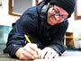 A man in a beanie and glasses leans over a piece of wood, sketching with a pencil. Wearing a dark jacket, his focus is evident as he works in a bright room with wooden furniture. This could mark the beginning of his next Brett Amory-inspired limited edition fine art giclee.