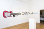 The gallery exhibits oversized hand-cut wood wall art: a red electric guitar, a black guitar, and a blue sword enhanced by acrylic art. Colorful icons add flair. Set against wooden floors and white walls with a central pedestal, it embodies Matt Ritchies artistic vision.