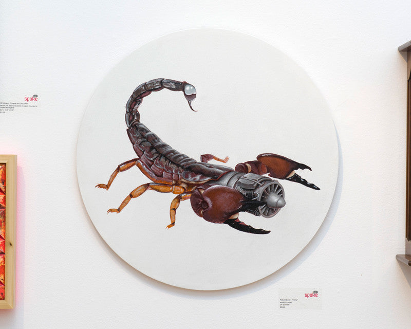 Robert Bowens circular artwork of a scorpion with mechanical elements, featuring gears and metallic parts, is displayed on Spoke Arts white wall. Accompanied by small labels, it evokes the ingenuity seen at events like the Supersonic Invitational.