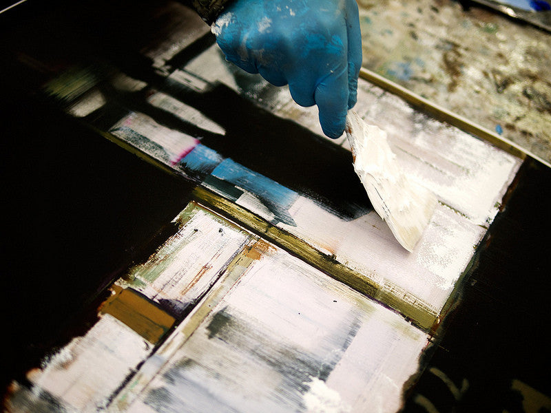 A gloved hand applies white paint with a palette knife to an abstract canvas featuring geometric shapes in black, white, and blue. The composition echoes Brett Amorys style, reminiscent of a fine art giclee from his limited edition series.