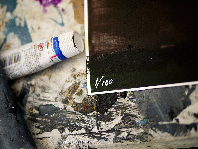 A paint-covered surface showcases a squeezed tube and partially visible artwork by Brett Amory. Marked 1/100 in white, it is the first piece in his fine art giclee limited edition series.