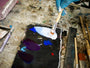 Blue-gloved hands mix white paint with blue and purple on a messy palette, surrounded by brushes and tools reminiscent of Brett Amorys studio for crafting limited edition fine art giclee prints.
