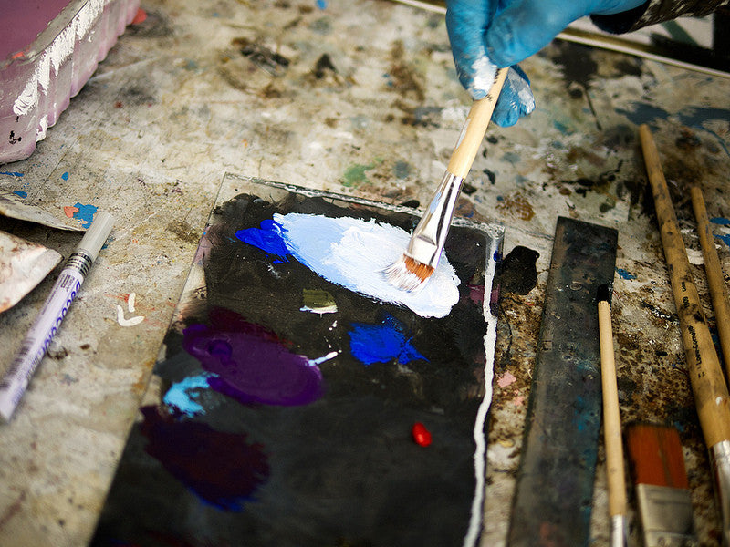 Blue-gloved hands mix white paint with blue and purple on a messy palette, surrounded by brushes and tools reminiscent of Brett Amorys studio for crafting limited edition fine art giclee prints.