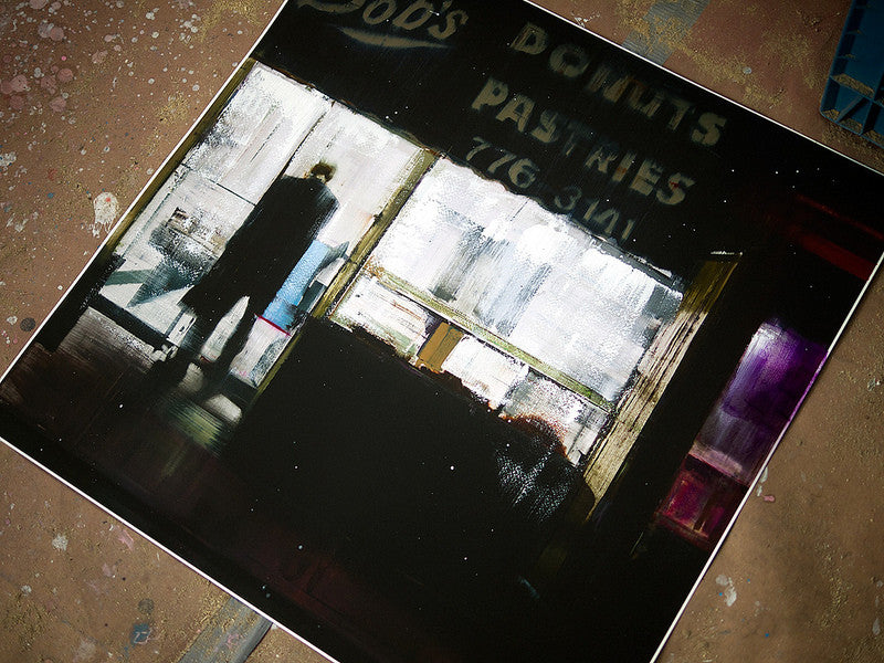 This limited edition fine art giclee by Brett Amory portrays a moody darkened shop interior, featuring a silhouette by the lit window gazing outside, with Donuts and Pastries signs partially illuminated.