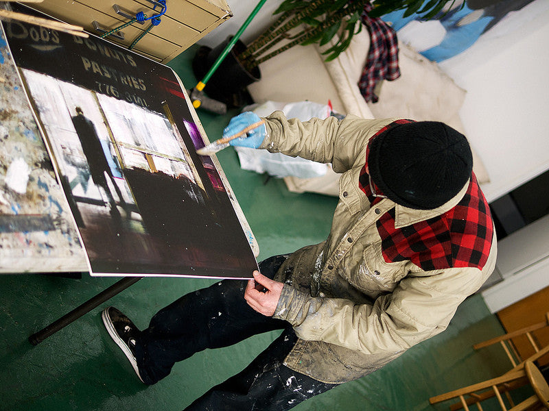 In a studio filled with art supplies and furniture, a person in a plaid-lined jacket and beanie paints on a canvas, capturing a silhouetted figure in a window. The scene echoes the vibe of Brett Amorys fine art giclee, suggesting the creation of a limited edition masterpiece.