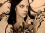A monochromatic portrait by Alessandra Maria features a young person with dark hair surrounded by lilies, gazing seriously to the side. The composition is enhanced with hints of gold leaf against a light background, creating a striking contrast reminiscent of her style.