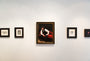 Explore the Chimerical exhibition on our minimalist gallery wall with five framed artworks. At the center is a striking Karla Ortiz portrait of a man on a red sofa, inspired by La Fae. Smaller black-and-white images are perfectly spaced against a plain white backdrop.