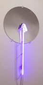 A round mirror on a white wall reflects the mesmerizing purple glow of Meryl Patakys glass neon art, featuring beautifully curved tubes.
