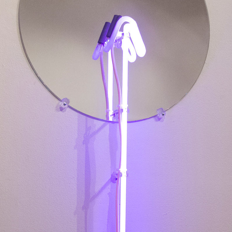 A round mirror on a white wall reflects the mesmerizing purple glow of Meryl Patakys glass neon art, featuring beautifully curved tubes.
