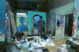 In a cluttered art studio, someone paints a vivid portrait on canvas. Papers and paint cans are scattered, with walls adorned by Andrew Hems dynamic artwork, reflecting a creative atmosphere reminiscent of mixed media on panel techniques.
