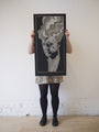 Against a plain white wall, a person holds up a Chuck Sperry screen print poster of a stylized black-and-white portrait with wavy hair. They’re dressed in a floral skirt and black tights, adding an artistic flair reminiscent of David Lynchs iconic aesthetic to the scene.
