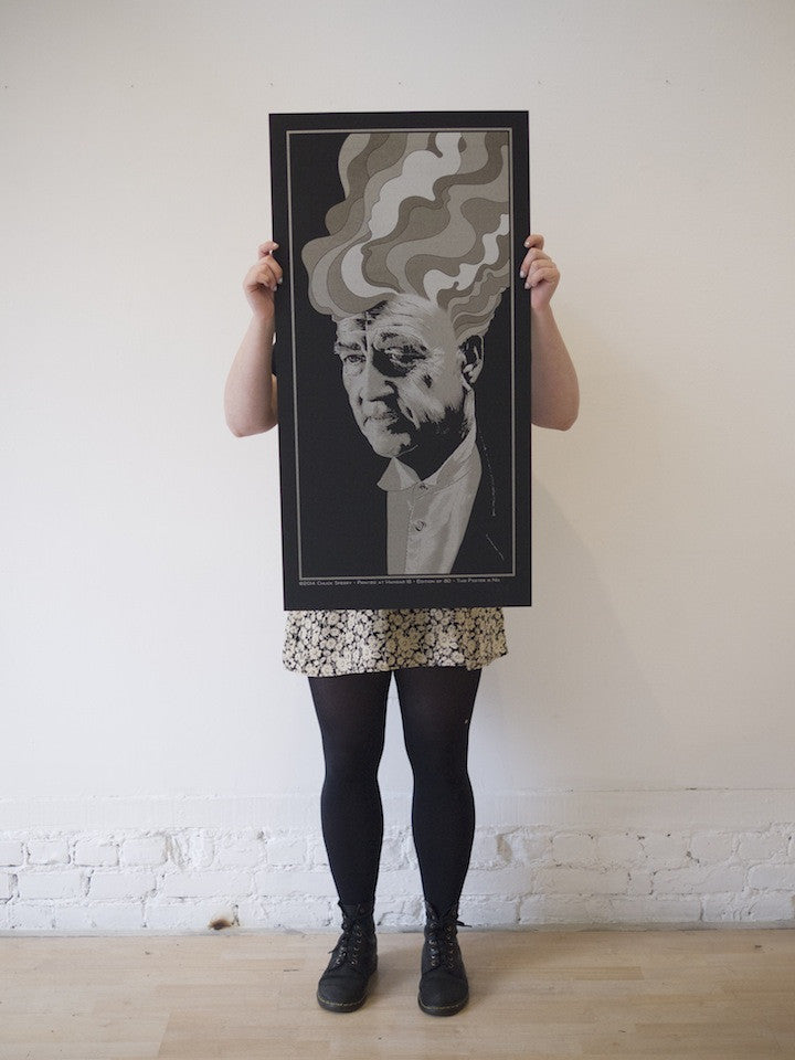 Against a plain white wall, a person holds up a Chuck Sperry screen print poster of a stylized black-and-white portrait with wavy hair. They’re dressed in a floral skirt and black tights, adding an artistic flair reminiscent of David Lynchs iconic aesthetic to the scene.