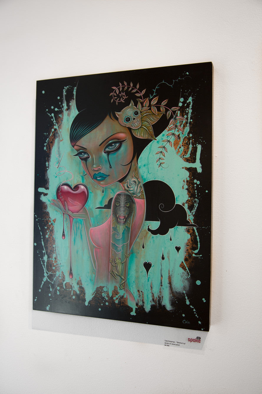 A Caia Koopman painting showcases a stylized female with dark hair, dramatic makeup, small skull and leaves in her hair. Acrylic on wood panel, it features a dripping heart and abstract patterns on a stitched dark and vibrant background.