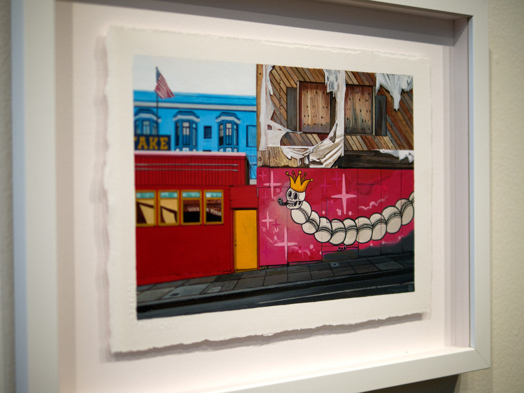 This custom framed artwork by Jessica Hess showcases a whimsical caterpillar in a crown on a pink wall, with her vibrant gouache style reflected in the background of a red building with flags and a blue one with large windows.