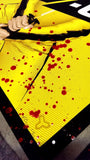 A silkscreen print poster by Tracie Ching showcases a yellow-haired warrior in vivid yellow gear with a katana, surrounded by red liquid splatters resembling blood. The mainly yellow backdrop is bordered in black and includes an abstract symbol in one corner.