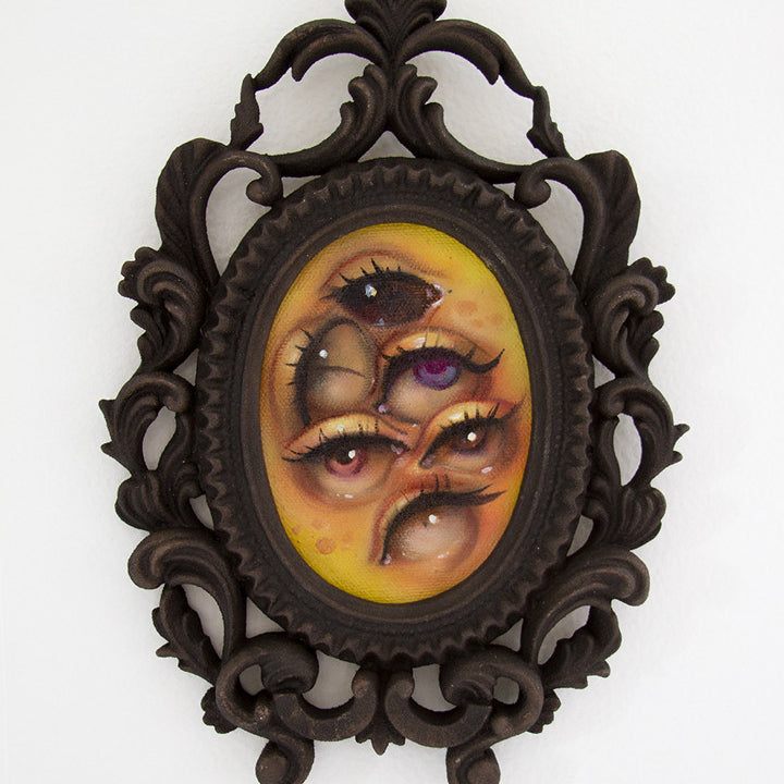 Tatiana Suarezs surreal oil on canvas painting is set in an ornate oval frame. It features a multi-eyed face with dark lashes and yellowish skin. The intricate scrollwork of the dark frame contrasts vividly, capturing the essence of the Through the Veil exhibition.