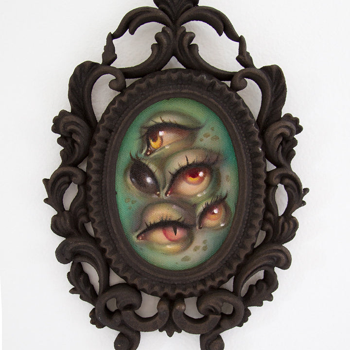 The ornate dark frame holds Tatiana Suarezs Through the Veil, an oil on canvas featuring multiple eyes in vibrant yellow and orange against a green textured background, creating an otherworldly, surreal effect with their varied sizes and orientations.