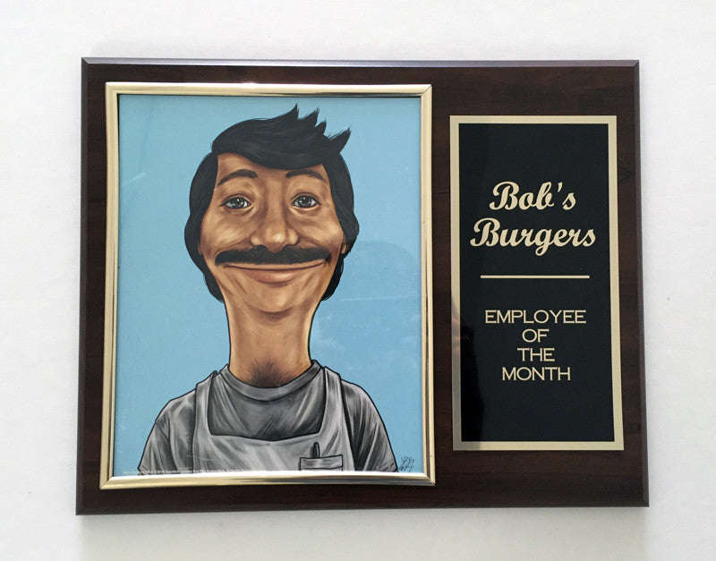 A framed Employee of the Month plaque by Cuyler Smith, featured at Spoke Art NYC, displays a caricature of a smiling man with a thick mustache on a blue background. Its wooden frame highlights the shows name and award title in gold lettering.