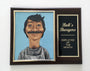A framed Employee of the Month plaque by Cuyler Smith, featured at Spoke Art NYC, displays a caricature of a smiling man with a thick mustache on a blue background. Its wooden frame highlights the shows name and award title in gold lettering.