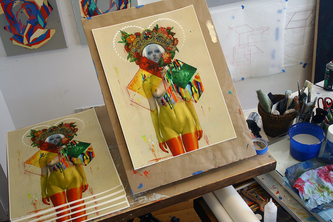 In the art studio, an eye-catching mixed-media piece by Erik Jones, featuring vivid colors and geometric designs, sits on the easel. A figure with a detailed headpiece is showcased. Limited edition giclee prints made with archival Epson HDR inks are displayed on the table.