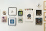 A gallery wall features Matt Ritchies cat-themed artworks, including vibrant acrylic paintings of colorful cats, small framed sketches, and ceramic cat faces. The original pieces are artfully arranged with a delightful touch of Lil Bubs influence in a mixed and visually appealing layout.