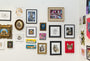 A gallery wall displays various framed cat artworks, including The Cat From Outer Space by Meghan Stratman. The collection creatively combines realistic, abstract, and whimsical depictions with diverse styles and colors on a white wall.