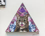 The triangular artwork by Johannah ODonnell features a central cat with big eyes and its tongue out, reminiscent of the Lil Bub exhibit in San Francisco. The background is filled with geometric patterns in purple and pink, flanked by smaller blue and pink cats, and crowned with a purple shape.