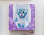 Displayed at the San Francisco gallery, this Johannah ODonnell acrylic painting shows a fluffy cat with blue paws and fur, playfully sticking its tongue out against a purple bokeh-style background. Part of the Lil Bub exhibit, it sits elegantly with a small plaque beneath it.