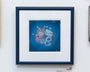 A framed artwork by Nicole Gustafsson showcases Lil Bubs face among flowers on a blue background. It features a black frame with a white mat, making it the ideal addition to any art exhibit.