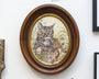 Displayed on a white wall, a round wooden frame by Jeremy Hush features an illustration of a green-eyed kitten playfully peeking through lush grass with its mouth open, all set against a light earthy background, reminiscent of the Lil Bub art exhibit.