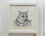 A sleek white-framed artwork by John Casey features a playful tabby cat with big green eyes and its tongue out, capturing the essence of the Lil Bub show. The drawing showcases colored pencil and ink details of black and white striped fur on a light-colored wall.
