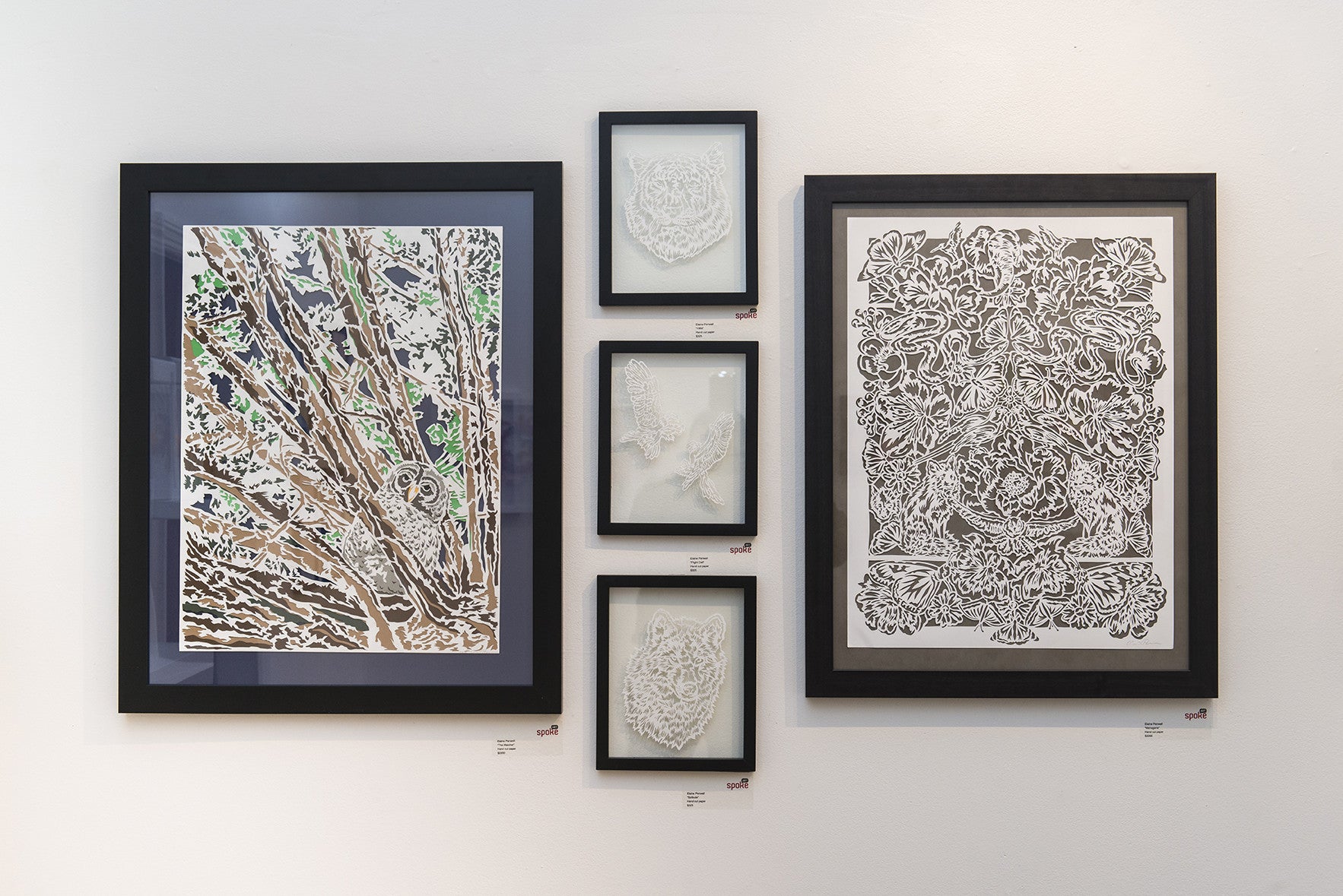 An art exhibition wall displays framed pieces: Left is Elaine Penwells vibrant owl in a tree; right is an intricate black and white floral and animal design inspired by 1984; center features three smaller textured white hand-cut paper artworks in diverse shapes.