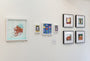 A gallery wall features a variety of framed artworks, including an octopus illustration and abstract shapes. A Mushroom Village printed giclee by Crowded Teeth adds charm to the display, while the neatly arranged frames on the white wall create an eclectic showcase.