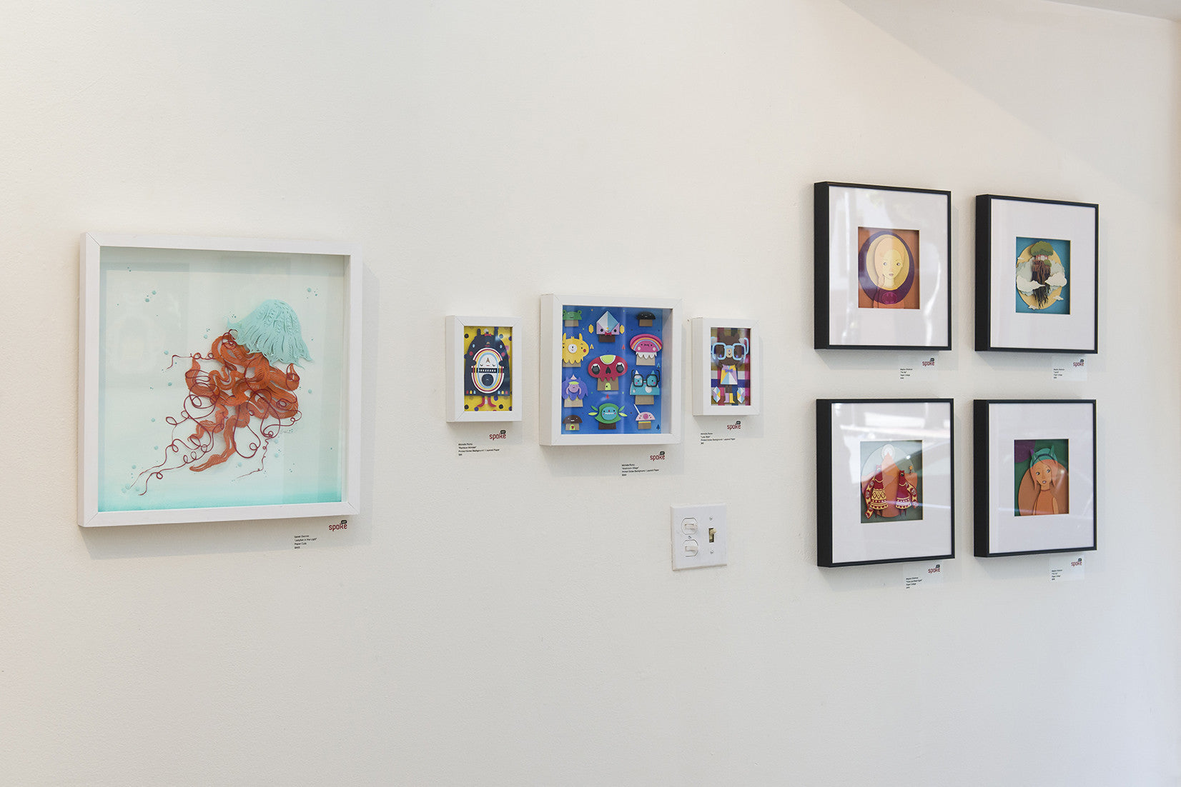 A white gallery wall showcases framed Crowded Teeth-inspired artworks, featuring whimsical and colorful illustrations. The frames, arranged in two rows, create a diverse visual experience that enhances each printed giclees vibrancy.