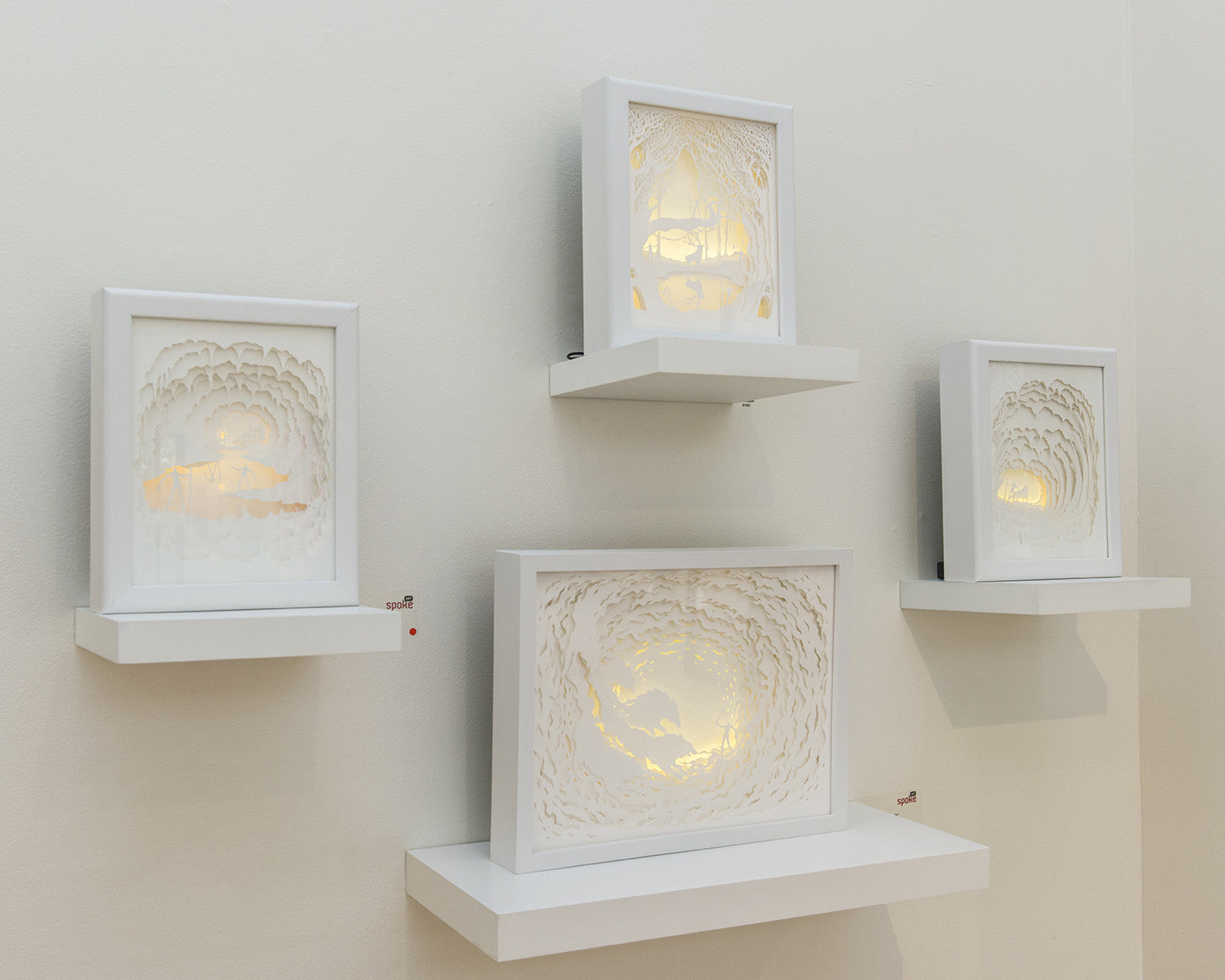 Four illuminated paper sculptures by Hari & Deepti are showcased on white shelves, each featuring intricate designs of tunnels or caves with a warm internal glow. The pieces vary in size and orientation.