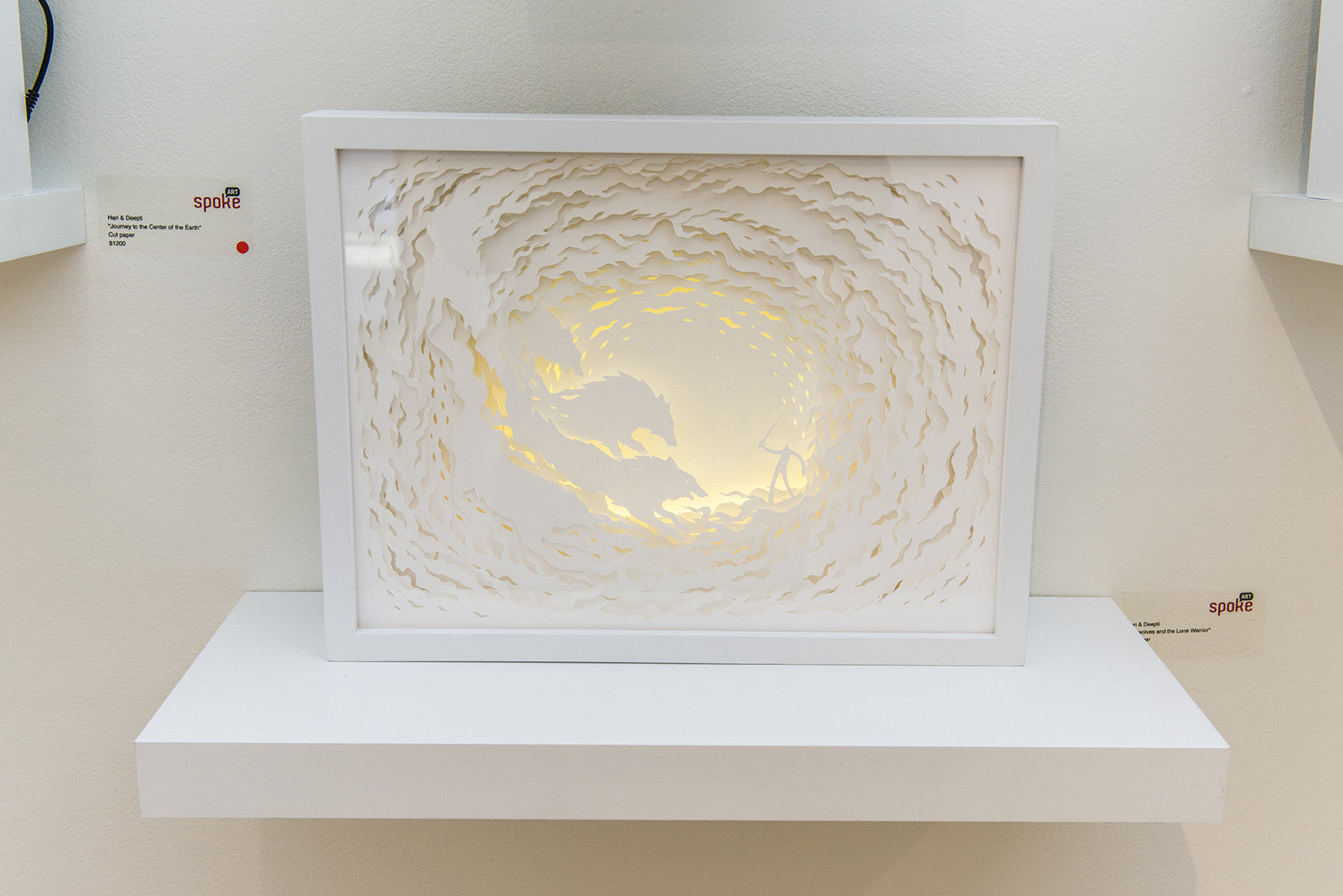 Hari & Deeptis layered paper art in a white frame showcases Firewolves and the Lone Warrior, featuring intricate wave cutouts and a creature confronting a figure. Soft lighting enhances the design, casting subtle shadows on a white shelf.