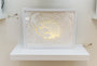 Hari & Deeptis layered paper art in a white frame showcases Firewolves and the Lone Warrior, featuring intricate wave cutouts and a creature confronting a figure. Soft lighting enhances the design, casting subtle shadows on a white shelf.
