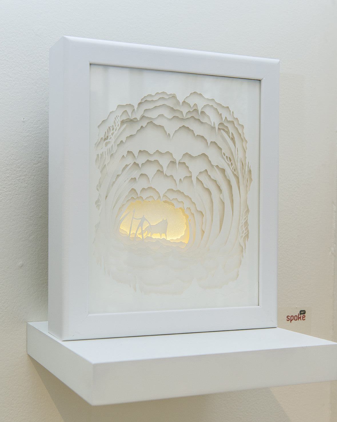 Hari & Deeptis white, layered paper artwork in a shadow box frame features a fox in a forest. Backlighting enhances the intricate design with subtle shadows. Evoking Where the Giants Rest, it sits elegantly on a white shelf against a light wall.