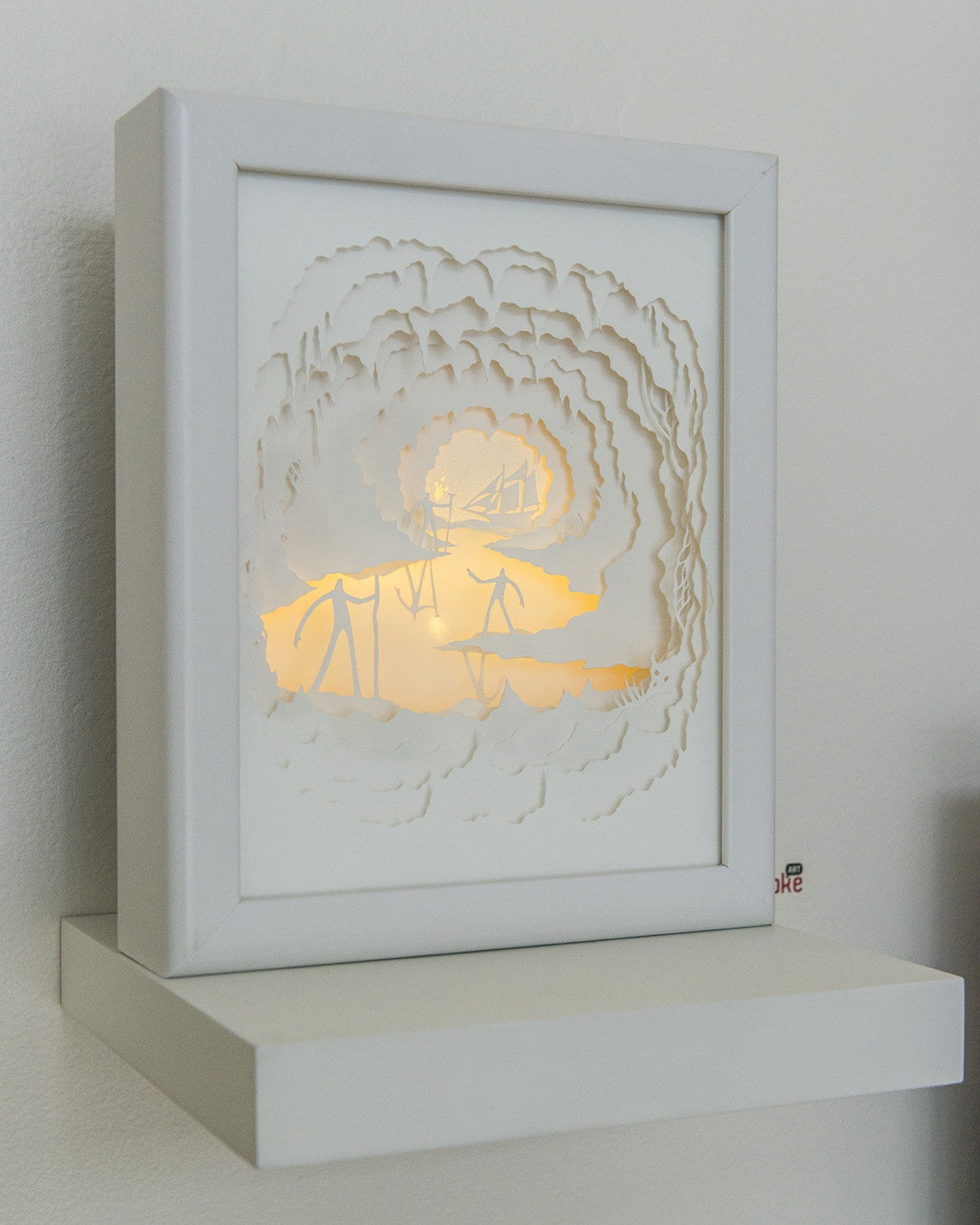 Hari & Deeptis white shadow box showcases intricate cut paper art with a layered landscape and figures. Backlit, it casts a warm glow highlighting the details, and sits on a white shelf against a light wall, evoking Journey to the Center of the Earth.