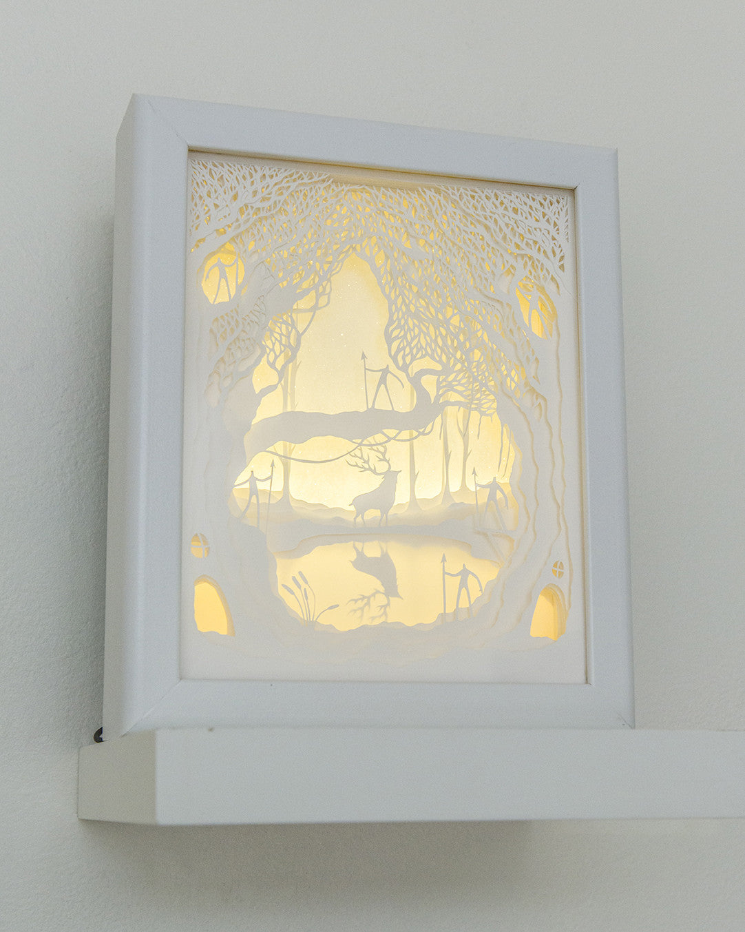 A white, intricately detailed cut paper lightbox from Hari & Deepti sits on a shelf. It features a silhouetted deer in an enchanted forest with trees and branches, softly illuminated to create a delicate and serene ambiance.